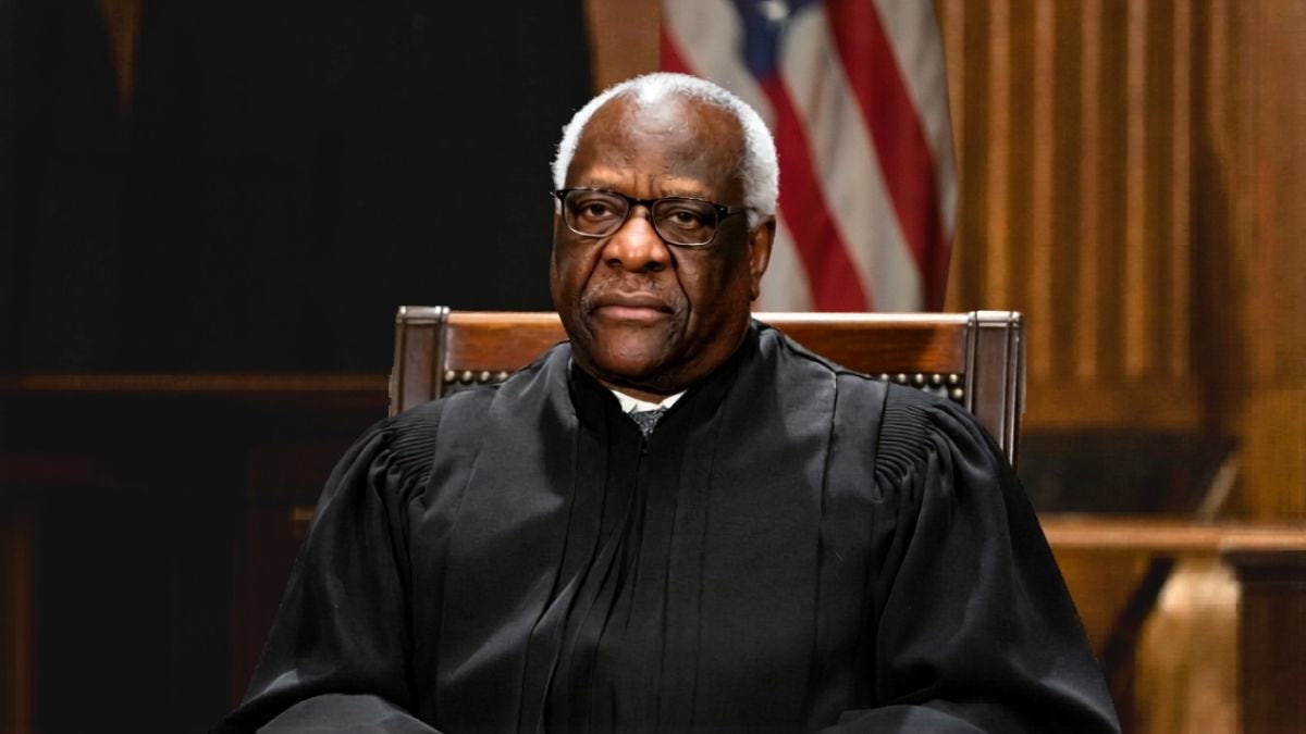 An older man in a black judicial robe and glasses sits in a wooden chair in a courtroom, with an American flag behind him.